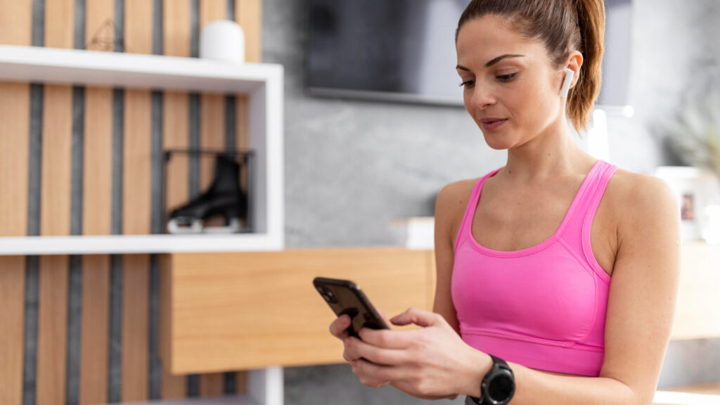 Deportista consultando app de fitness en smartphone mientras usa smartwatch negro y top deportivo rosa, con auriculares inalámbricos blancos, en ambiente interior con estantería de madera