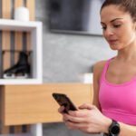 Deportista consultando app de fitness en smartphone mientras usa smartwatch negro y top deportivo rosa, con auriculares inalámbricos blancos, en ambiente interior con estantería de madera