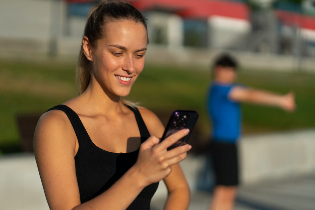 Deportista consultando datos de entrenamiento entre smartwatch y smartphone durante descanso en escaleras, usando top negro deportivo, auriculares inalámbricos blancos y bolsa deportiva con detalles neón