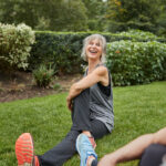 Mujer sonriente realizando estiramientos al aire libre en un parque, destacando los beneficios del ejercicio para la salud mental.