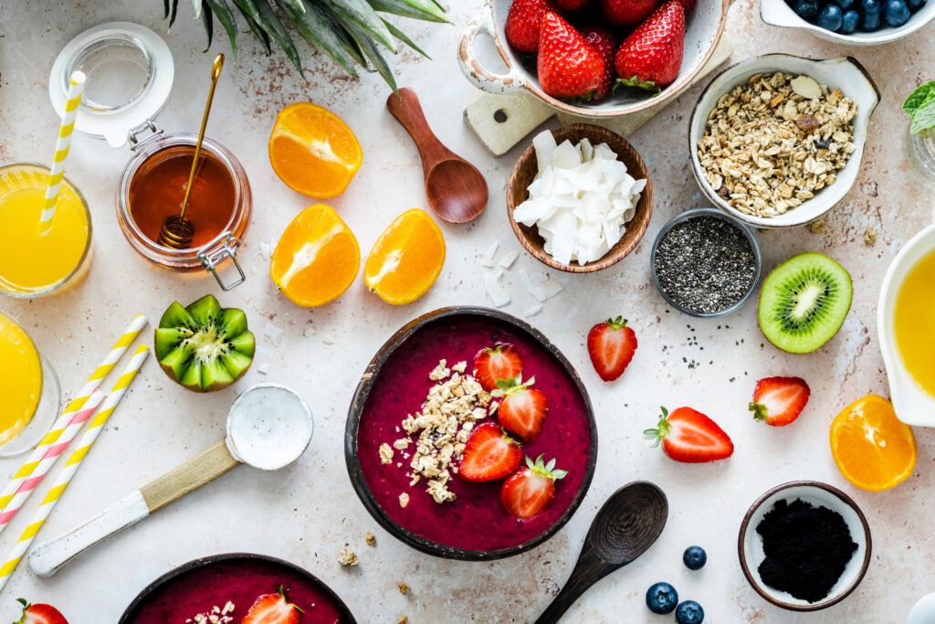 Smoothie bowl de color morado intenso decorado con fresas frescas y granola, rodeado de ingredientes saludables como naranjas, kiwi, miel, semillas de chía, coco rallado, arándanos y zumo de naranja con pajitas rayadas