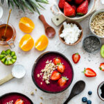 Smoothie bowl de color morado intenso decorado con fresas frescas y granola, rodeado de ingredientes saludables como naranjas, kiwi, miel, semillas de chía, coco rallado, arándanos y zumo de naranja con pajitas rayadas
