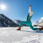 Persona realizando postura de yoga con brazos elevados, vistiendo conjunto térmico turquesa y botas de nieve marrones, sobre pista nevada con montañas y pinos nevados durante el invierno
