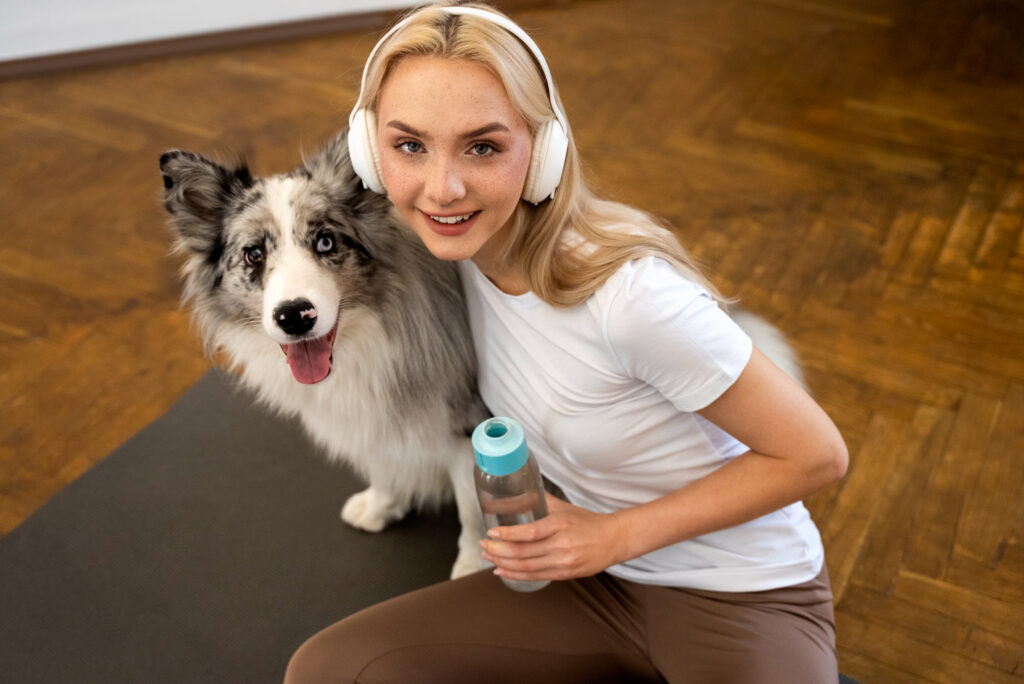 Persona en ropa deportiva blanca y auriculares blancos sobre esterilla negra, posando junto a su Border Collie gris y blanco, sosteniendo botella de agua con tapa turquesa, sobre suelo de madera