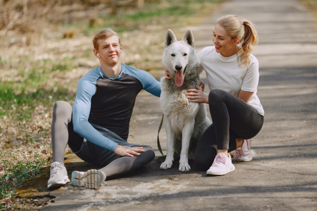 Pareja deportiva descansando durante entrenamiento al aire libre junto a su perro husky blanco, vistiendo ropa deportiva en azul y blanco, y zapatillas deportivas en tonos claros sobre sendero natural