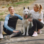 Pareja deportiva descansando durante entrenamiento al aire libre junto a su perro husky blanco, vistiendo ropa deportiva en azul y blanco, y zapatillas deportivas en tonos claros sobre sendero natural