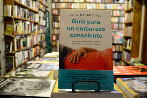 Libro "Guía para un embarazo consciente" de Laia Casadevall expuesto en estantería de librería, mostrando portada con vientre embarazado en rojo y título en turquesa, rodeado de otros libros en fondo desenfocado.