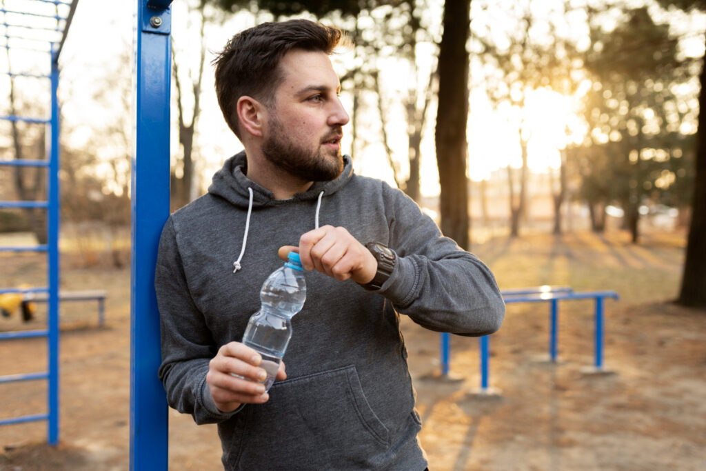 Hombre joven hidratándose con una botella de agua durante su entrenamiento al aire libre en un parque al atardecer.