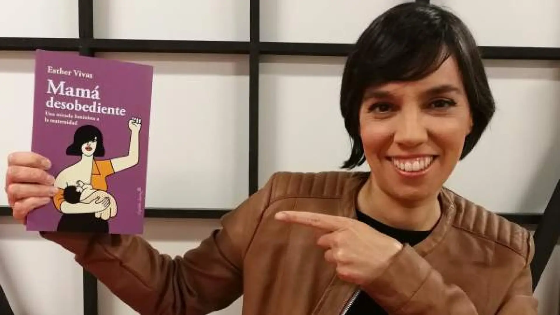 Presentadora sonriente con chaqueta marrón señalando la portada del libro "Mamá desobediente" de Esther Vivas, que muestra una ilustración de mujer amamantando con puño en alto sobre fondo morado