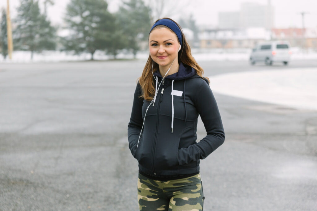 Persona con sudadera negra con capucha, cinta deportiva azul y auriculares blancos, realizando calentamiento en exterior nevado con árboles de fondo y pantalones de camuflaje