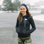Persona con sudadera negra con capucha, cinta deportiva azul y auriculares blancos, realizando calentamiento en exterior nevado con árboles de fondo y pantalones de camuflaje