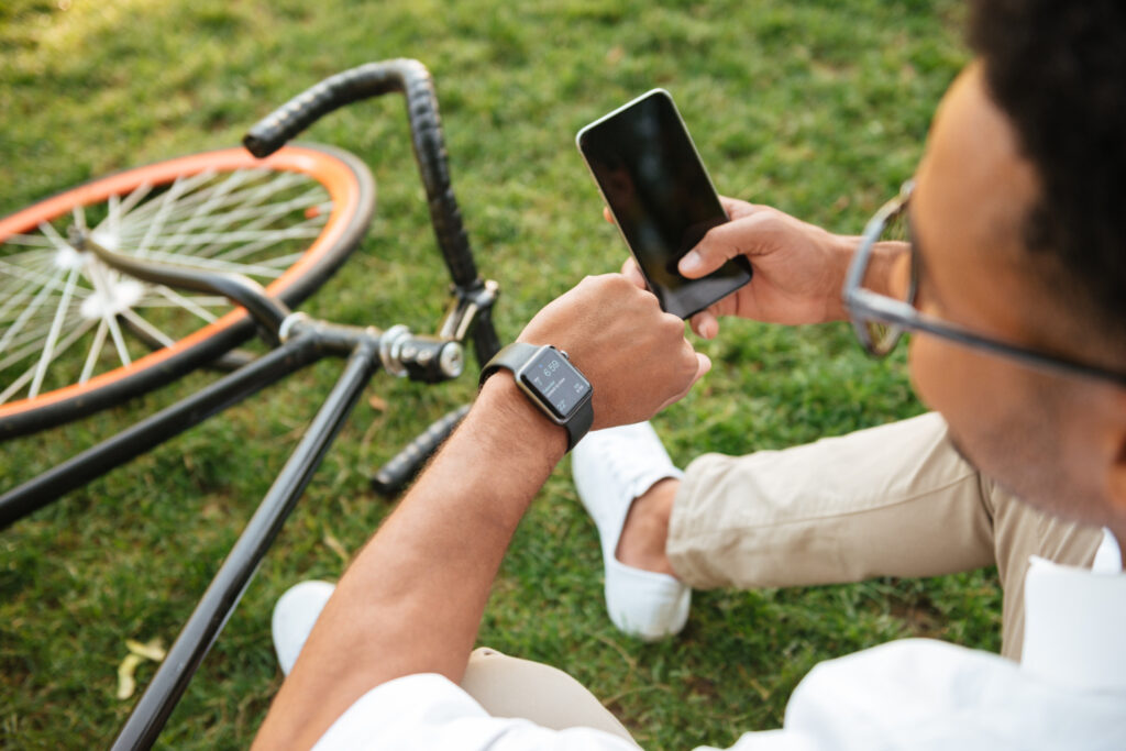 Ciclista sentado en césped configurando su smartwatch y smartphone junto a bicicleta con rueda naranja, vistiendo ropa deportiva clara y gafas, durante pausa de entrenamiento