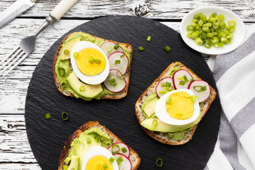 Tres tostadas de pan integral con aguacate machacado, huevo cocido, rodajas de rábano y cebollino picado, servidas sobre plato de pizarra negra con guarnición de cebollino fresco en bol blanco