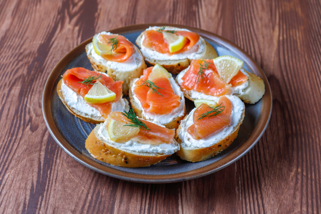 Tostadas gourmet con salmón ahumado y queso crema sobre plato gris oscuro, decoradas con rodajas de limón y eneldo fresco, servidas sobre mesa de madera rústica