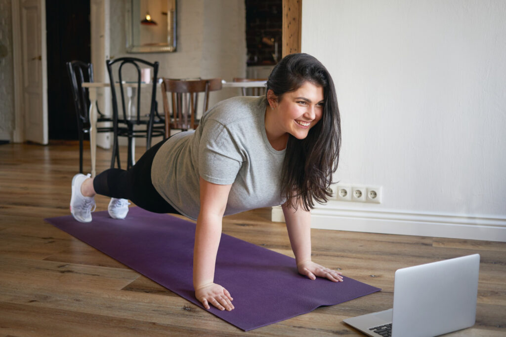 Mujer sonriente realizando ejercicio de plancha sobre esterilla morada, vistiendo camiseta gris y leggings negros, siguiendo rutina en laptop, en espacio luminoso con suelo de madera y sillas de café como fondo