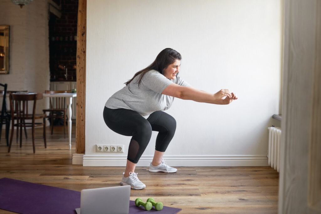 mujer sonriente realizando sentadilla con brazos extendidos, vistiendo camiseta gris y leggings negros, siguiendo rutina en laptop junto a mancuernas verdes sobre esterilla morada, en espacio luminoso con suelo de madera y sillas de café como fondo