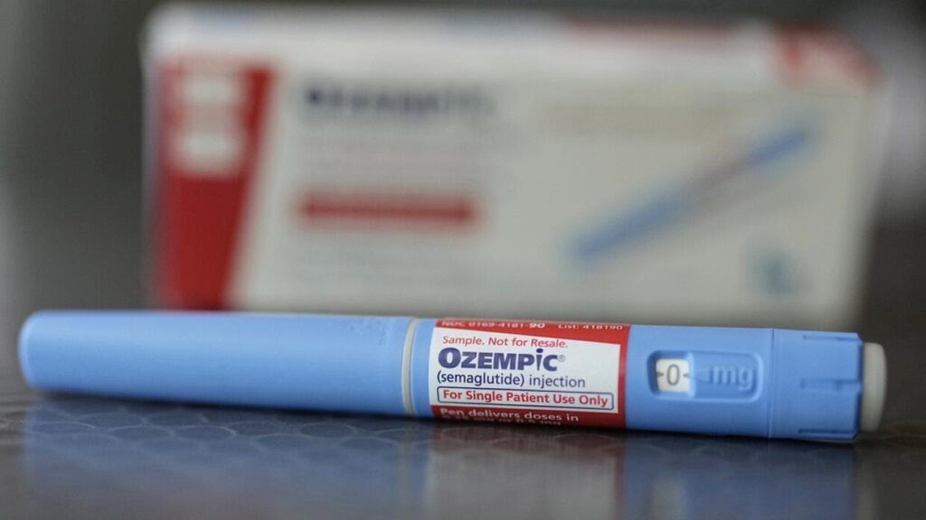 Pluma inyectable Ozempic de semaglutida en color azul claro con etiqueta roja de muestra, dosis de 0 mg, sobre superficie oscura con caja de medicamento desenfocada en segundo plano