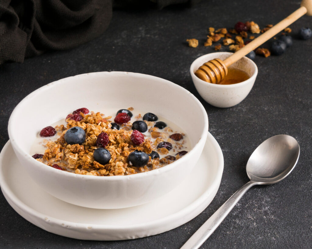 Bowl blanco con yogur proteico, granola casera crujiente, arándanos frescos y frambuesas sobre plato blanco redondo, acompañado de miel dorada en cuenco pequeño, fondo oscuro texturizado, receta saludable post entrenamiento